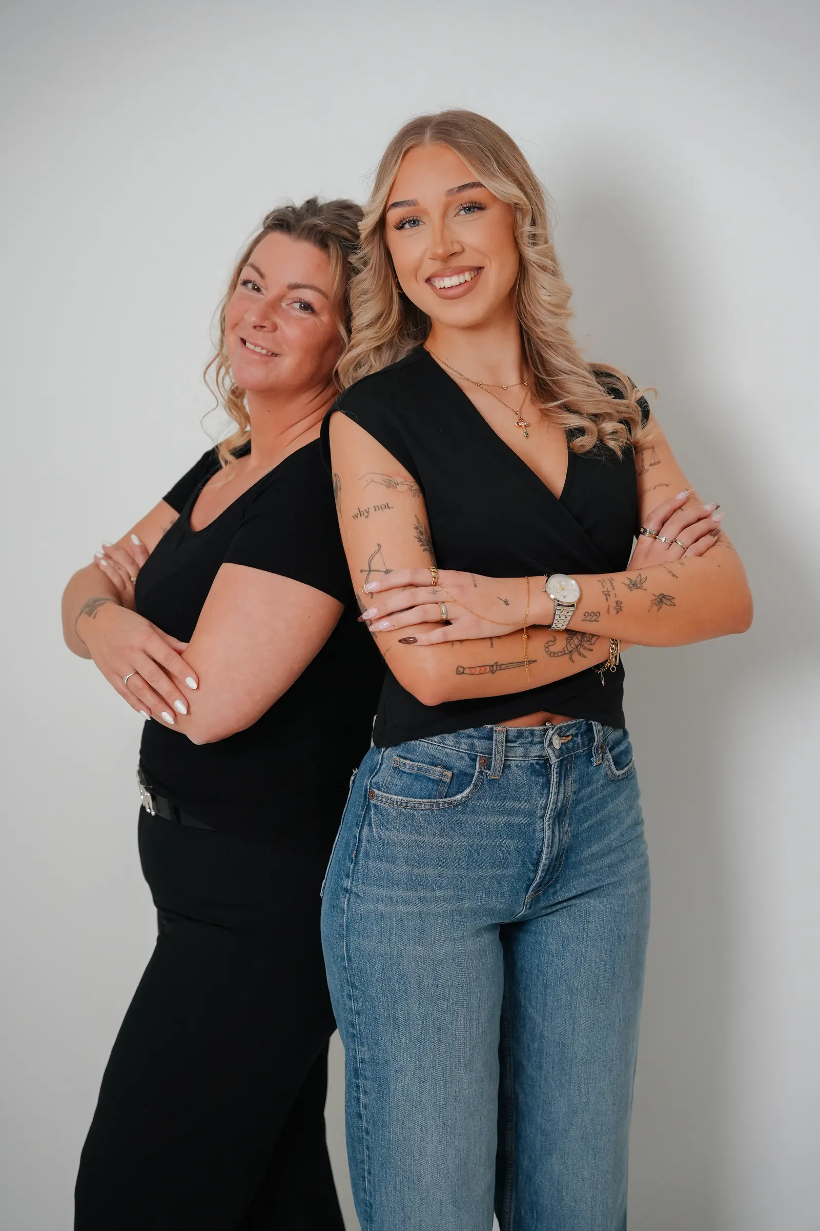 Diana und Jasmin stehen Rücken an Rücken mit verschränkten Armen vor einer hellen Wand